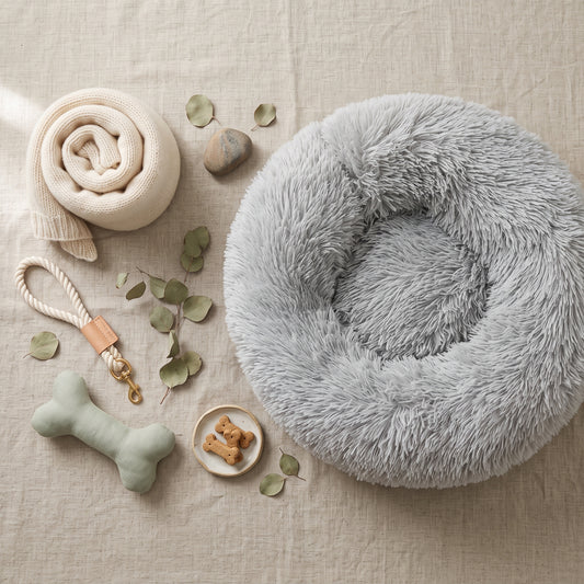 Calming Donut Pet Bed in gray plush faux fur, flat lay with a cream throw, rope toy, plush bone and dog treats